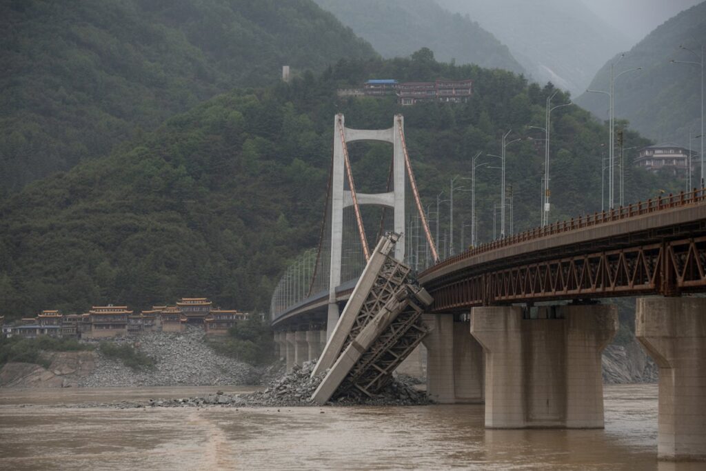 Wide view of the collapsed bridge in a mountainous valley, showing pier layout and surrounding terrain.