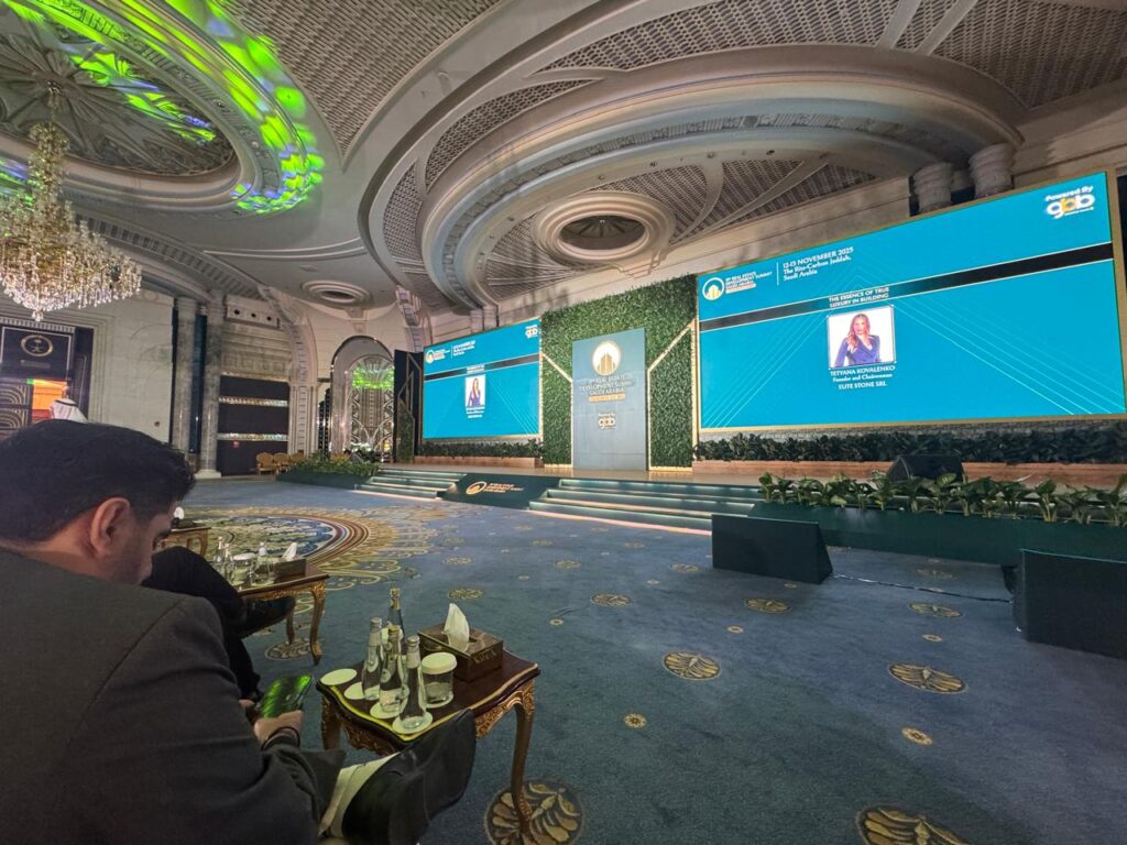 متحدثة على المسرح في قمة جدة العقارية، أمام جدران خضراء وشاشات تعرض "جوهر الفخامة الحقيقية في البناء