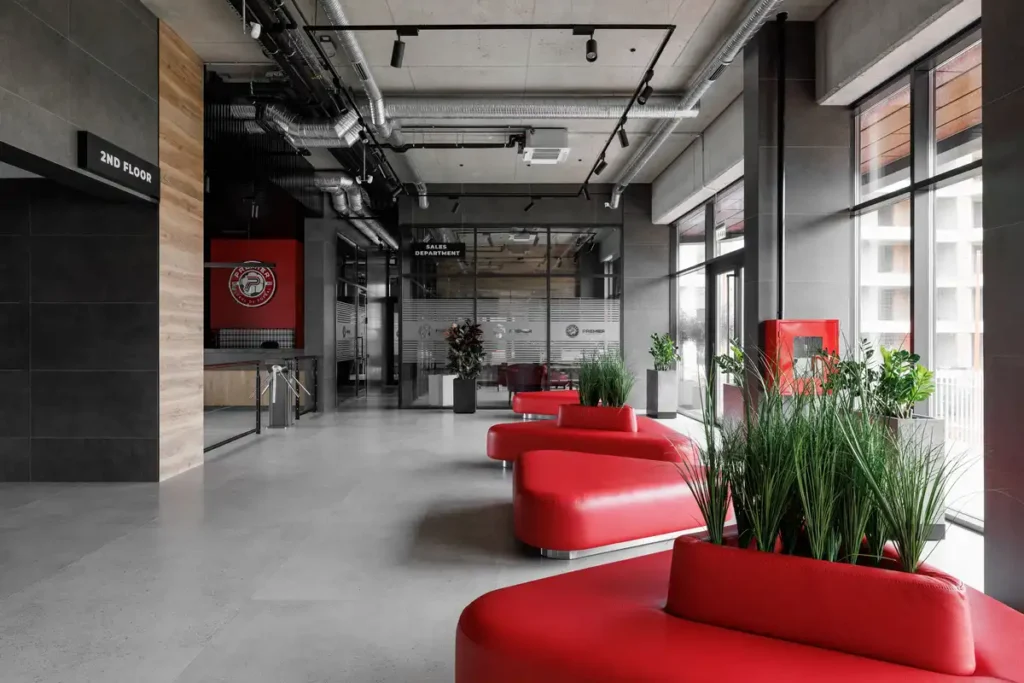 ردهة داخلية حديثة بمقاعد حمراء مريحة، وأرضيات رمادية، وسقف مكشوف يظهر أنابيب التهوية، مع نباتات خضراء تضفي لمسة طبيعية.