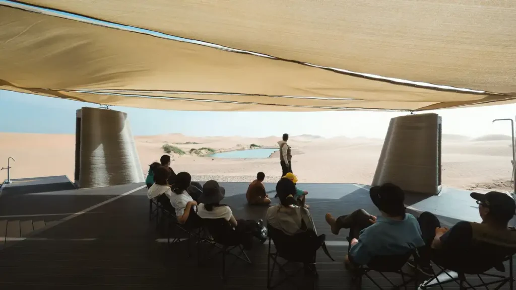 مجموعة من الزوار يستمتعون بإطلالة على الصحراء من تحت سقف قماشي في "ديزرت آرك"، محاطين بأعمدة خرسانية مطبوعة ثلاثيًا.