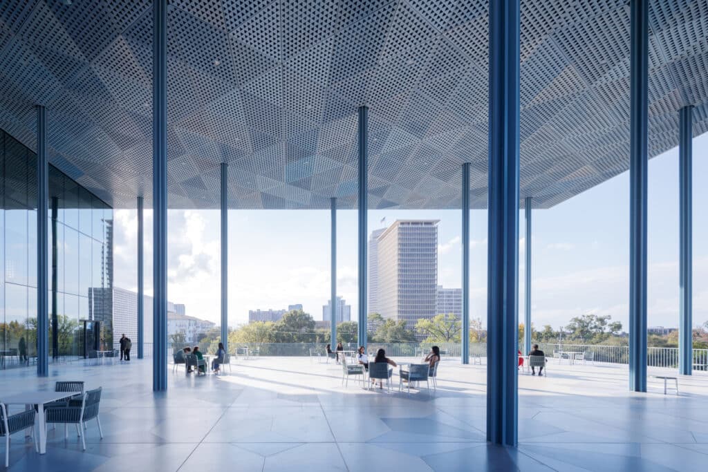 A shaded pavilion at the Ismaili Center, where modern architecture meets the urban skyline through a perforated ceiling and blue columns casting dappled light.