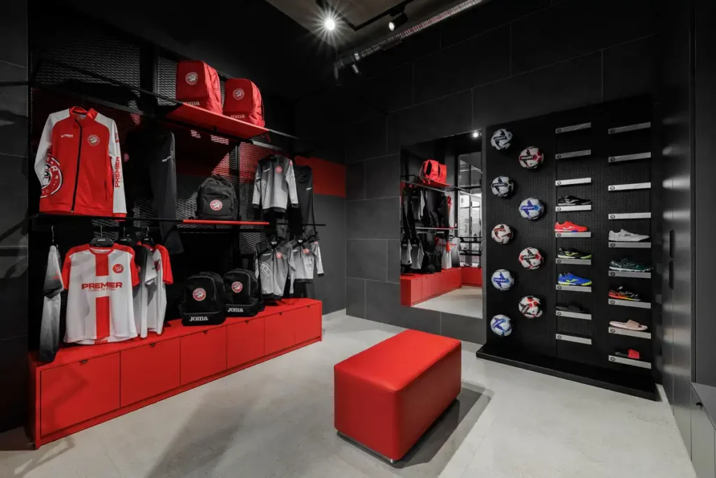 متجر رياضي داخلي بتصميم أسود وأحمر، يعرض ملابس وكرات وأحذية على أرفف مُنظمة، مع مقعد أحمر في المنتصف.