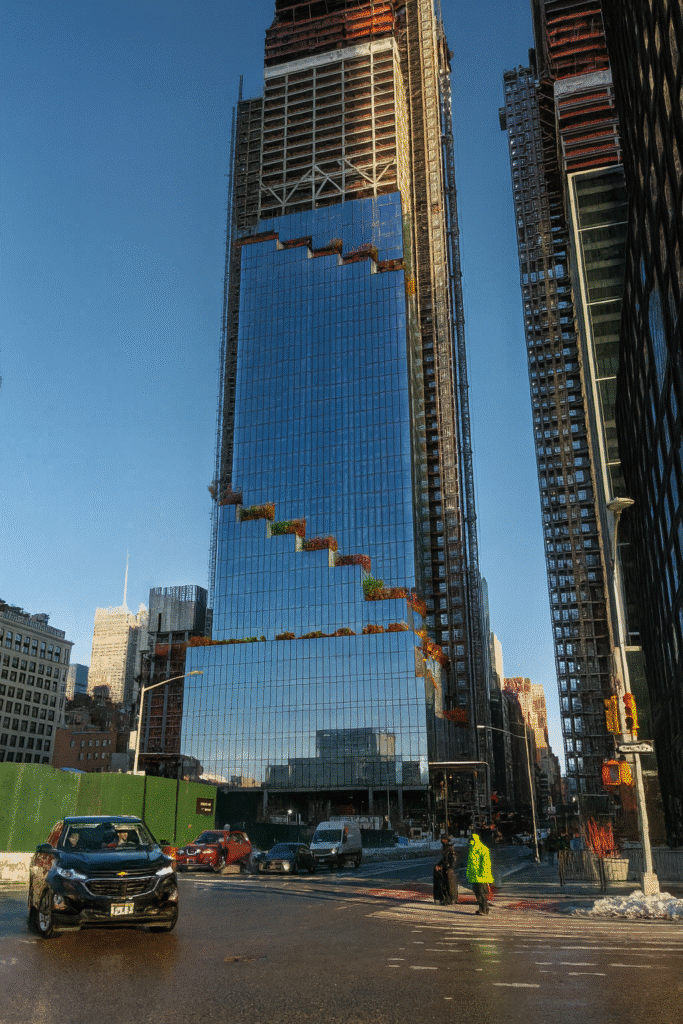 برج ذا سبايرل في نيويورك أثناء مرحلة البناء، بواجهته الزجاجية العاكسة وتصميمه المتدرج المميز، مع حركة مرور ومارة في الشارع أسفل المبنى.