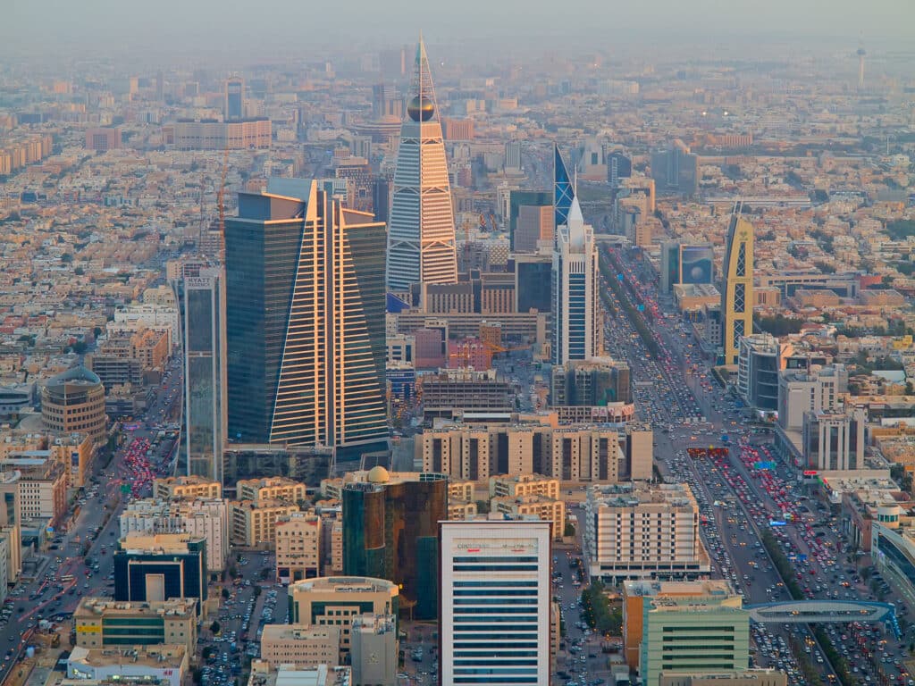A high-angle, panoramic view of Riyadh's modern downtown skyline at sunset, featuring prominent skyscrapers like the Kingdom Centre, flanked by busy multi-lane highways and dense urban development.
