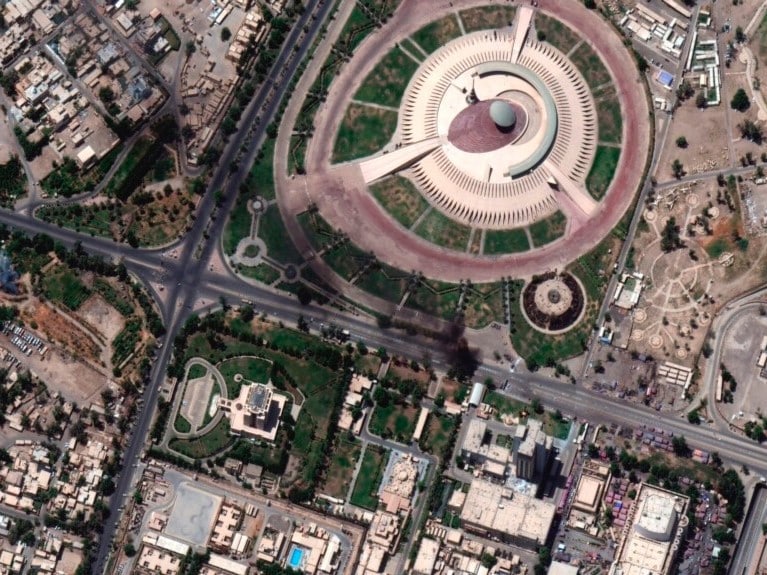 A top-down satellite view of the Monument of the Unknown Soldier in Baghdad, featuring its iconic circular plaza and inclined concrete shield design.