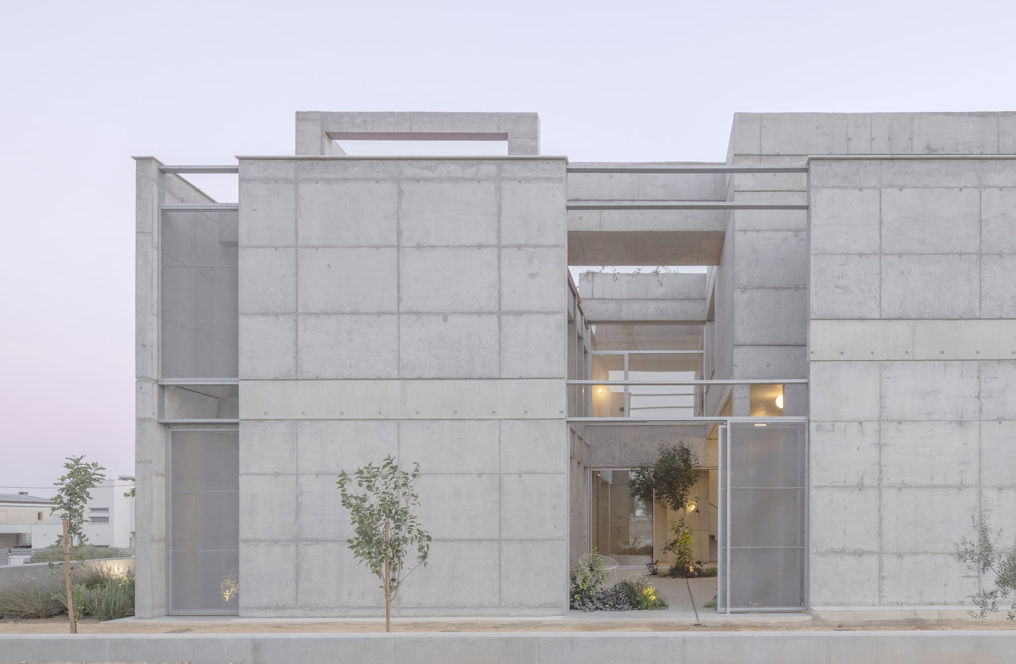 Main façade of the Carved Concrete house at dusk, showing solid masses intertwined with thin metal structural details and the illuminated entrance.