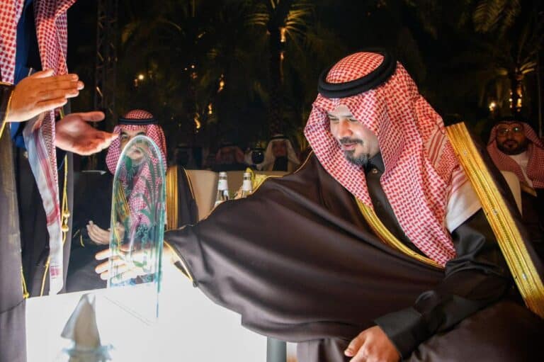 Saudi official extending hand toward an illuminated glass display during Al-Mustadill Orchard launch event in Medina, night, traditional attire.