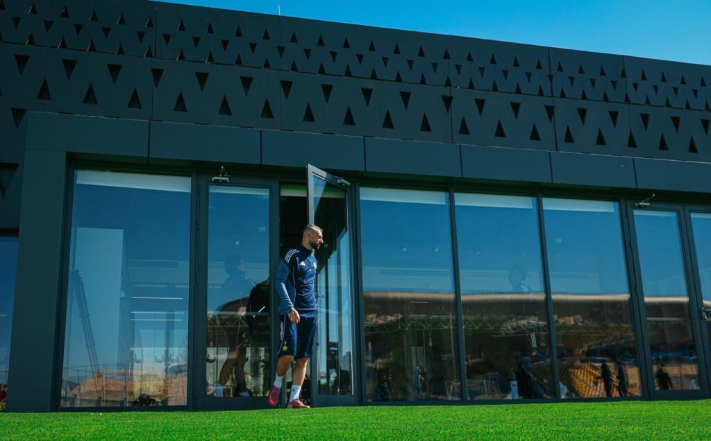 لاعب كرة قدم يخرج من منشأة تدريب حديثة ذات واجهة داكنة مثقبة وزجاجية، ليقف على العشب الصناعي تحت سماء صافية.