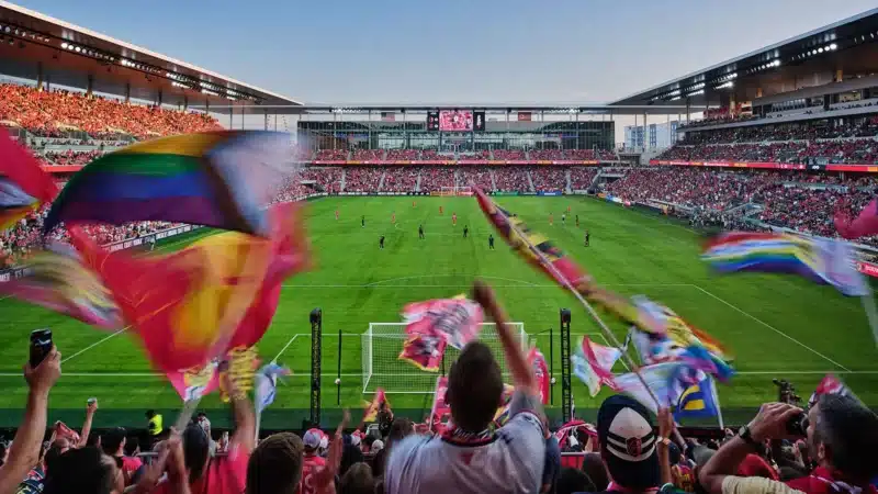 المشجعون يلوحون بالأعلام داخل الاستاد المندمج، يحيطون بالمستطيل الأخضر تحت إضاءة النهار.