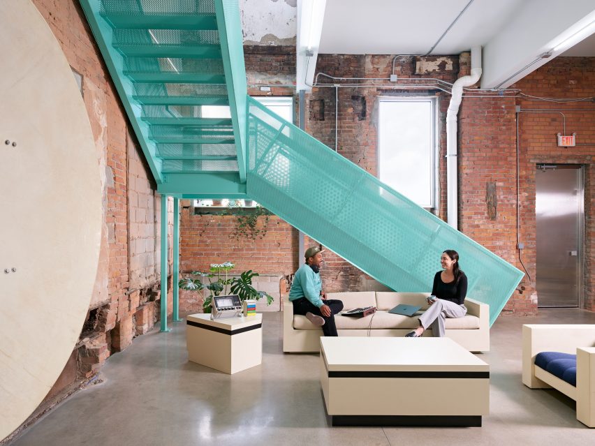 Two people seated on a modern beige sofa beneath a bold aqua perforated metal staircase, surrounded by exposed brick walls and industrial ductwork inside Assemble Sound’s studio.