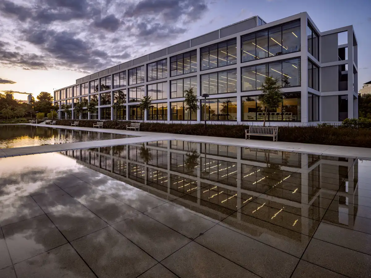 The sleek horizontal glass facade of the building reflecting the sunset and cloudy sky in a still water pond, emphasizing the beauty of the Sustainable Headquarters Design.