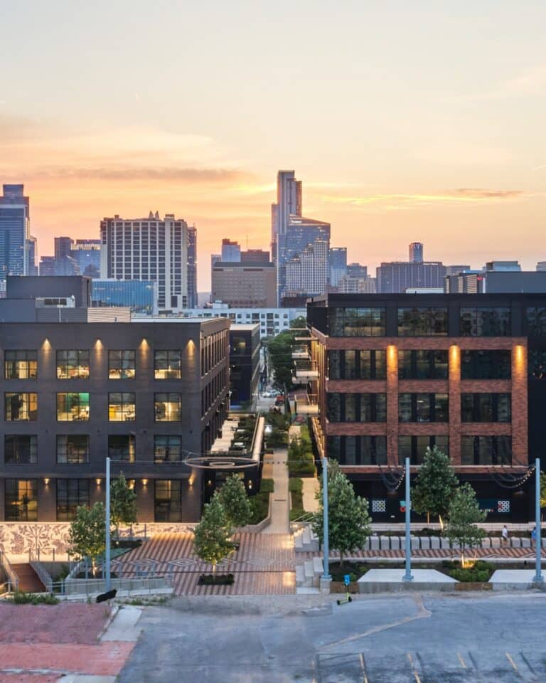 Centro West mixed-use development in East Austin at sunset, featuring two five-story brick buildings flanking a pedestrian alley with illuminated windows and the downtown skyline in the background.