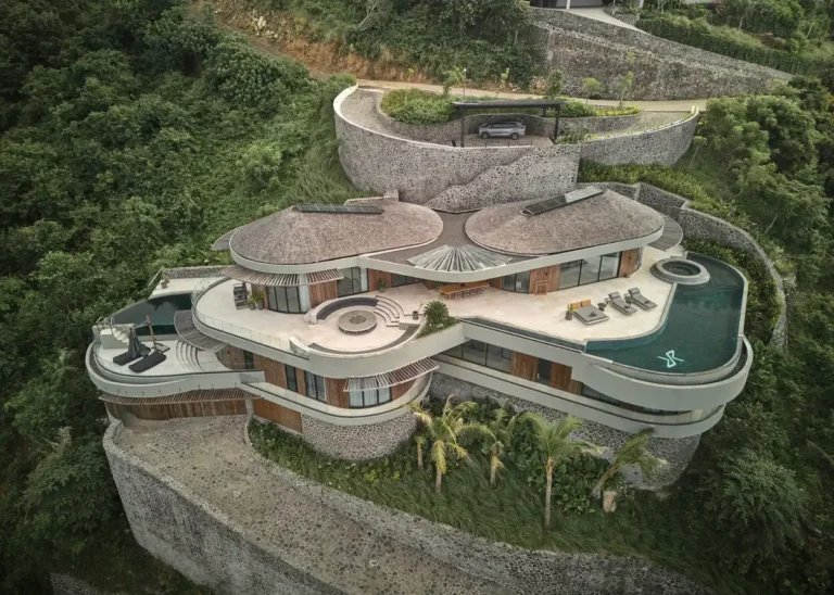 Aerial view of Nyrenstone Estate Lombok, a circular hillside villa with terraced stone retaining walls and organic pool shapes nestled in dense greenery.