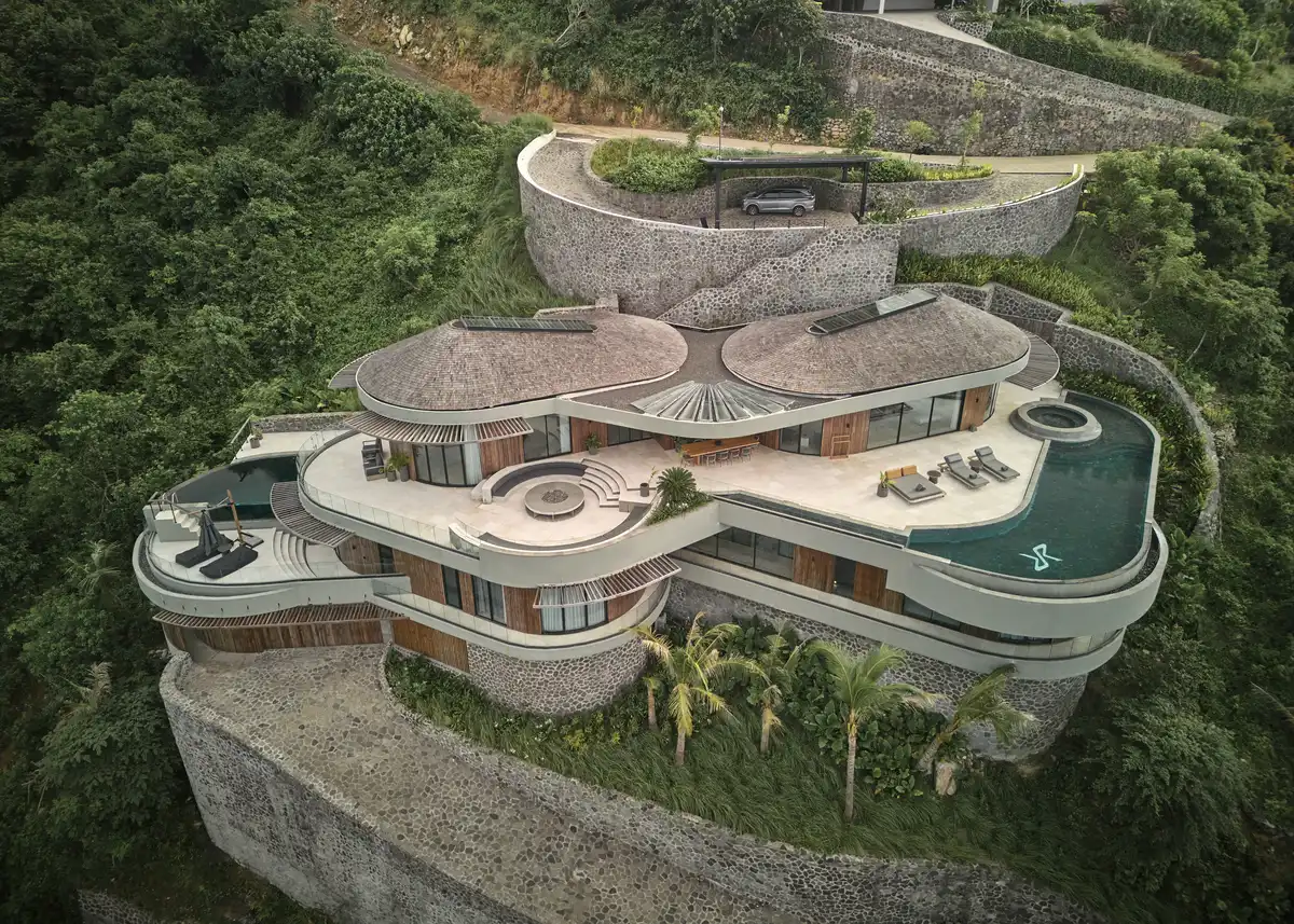 Aerial view of Nyrenstone Estate Lombok, a circular hillside villa with terraced stone retaining walls and organic pool shapes nestled in dense greenery.