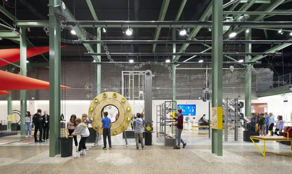 يتفاعل الأطفال والبالغون مع معروضات ميكانيكية ضخمة داخل قاعة عرض صناعية واسعة ذات أعمدة فولاذية خضراء مكشوفة وأرضيات خرسانية ملمعة.