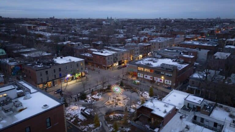Aerial view of Place des Fleurs-de-Macadam in Montreal at dusk, featuring the Un Brin d’Soleil light installation suspended above a snow-covered urban square surrounded by residential and commercial buildings.