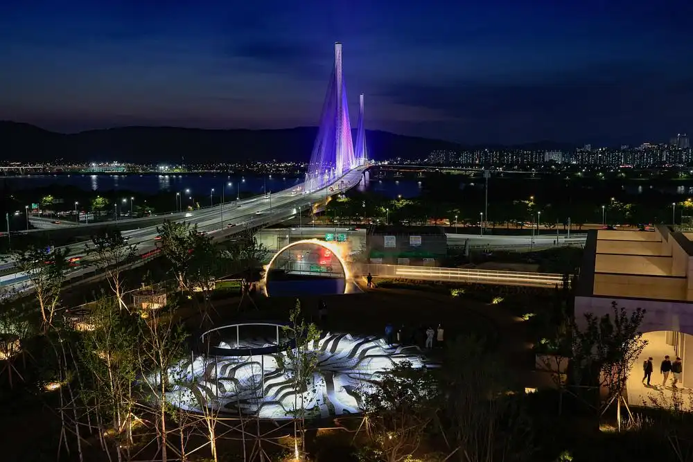 Night view of Arc ZERO: Eclipse Seoul installation on a rooftop garden, featuring a half-ring of mist and light reflected in water, with the illuminated Banpo Bridge in the background.