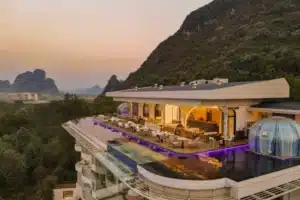 Aerial view of LS Hotel in Guilin at dusk, featuring a rooftop infinity pool with purple LED lighting, glass-walled lounge areas, and a transparent dome structure, all framed by lush karst mountains.