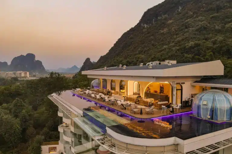Aerial view of LS Hotel in Guilin at dusk, featuring a rooftop infinity pool with purple LED lighting, glass-walled lounge areas, and a transparent dome structure, all framed by lush karst mountains.