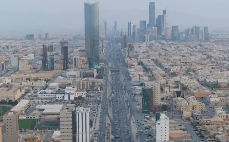 An aerial, high-angle view of a multi-lane highway (King Fahd Road) cutting through the center of Riyadh, flanked by modern skyscrapers and dense urban buildings under a hazy sky.