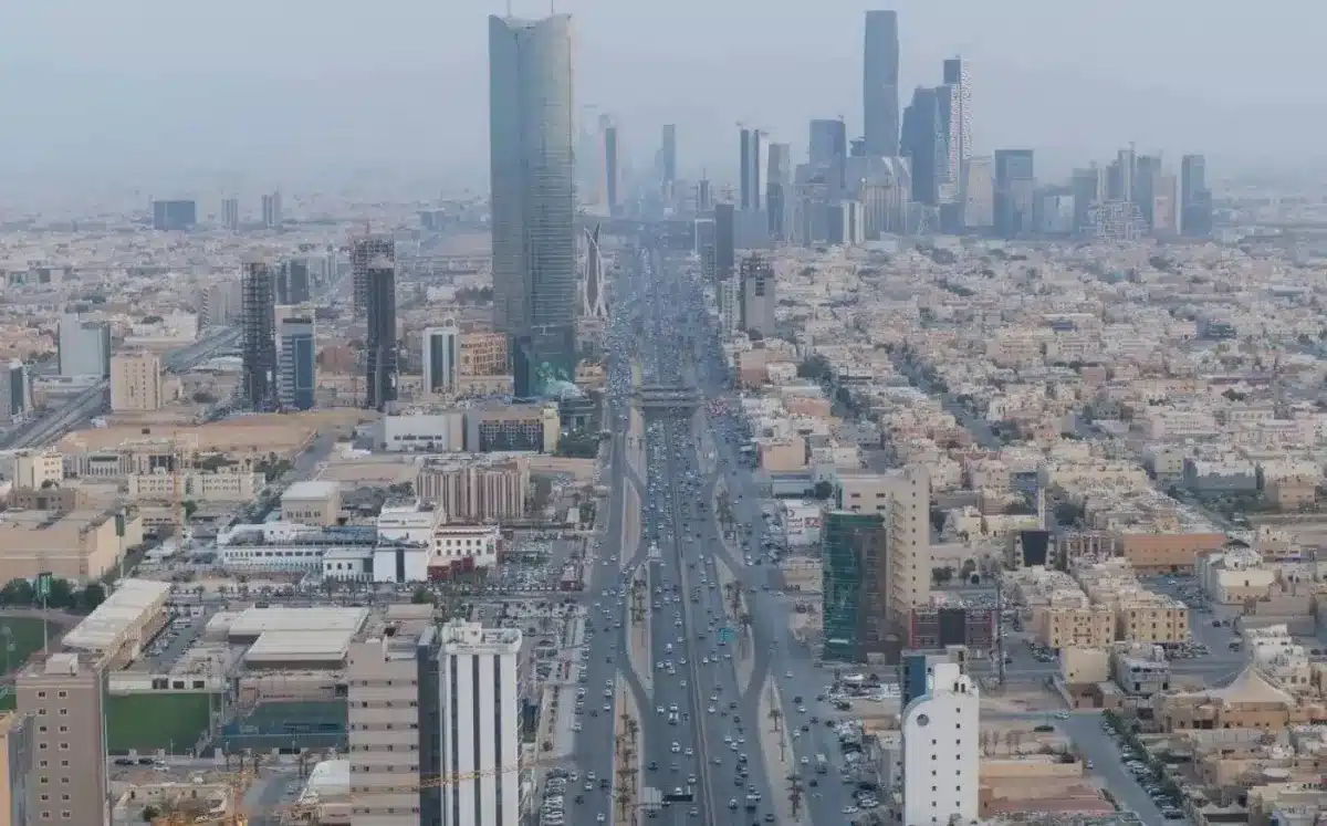 An aerial, high-angle view of a multi-lane highway (King Fahd Road) cutting through the center of Riyadh, flanked by modern skyscrapers and dense urban buildings under a hazy sky.