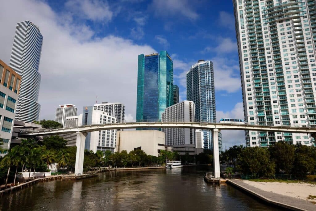 A view of a city river lined with modern glass skyscrapers, featuring a white elevated transit bridge with a blue and white train crossing over the water.