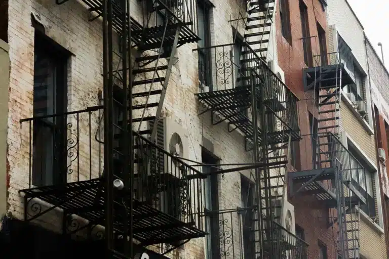 A close-up, low-angle shot of weathered brick apartment buildings featuring zigzagging metal fire escapes, decorative railings, and window-mounted air conditioning units.