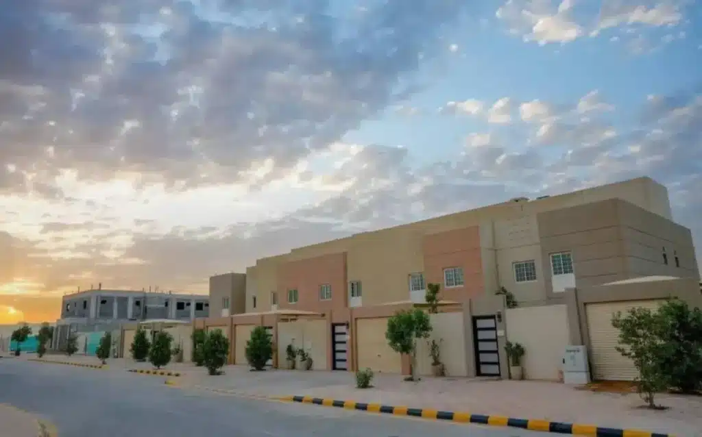 A row of modern two-story townhouses with beige and tan facades under a cloudy sunset sky in a quiet Saudi Arabian neighborhood.