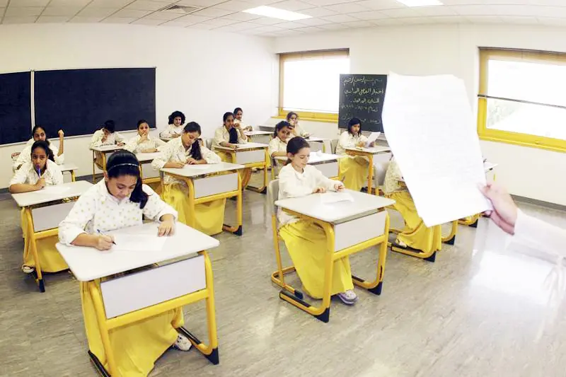 A group of female students in school uniforms sitting at individual desks and writing on exam papers in a brightly lit classroom.