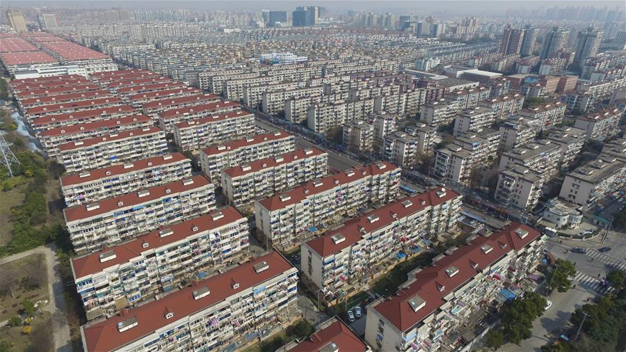 An aerial, wide-angle shot of a massive, dense residential area with rows upon rows of identical, multi-story apartment buildings with red-tiled roofs, stretching into the distance under a hazy sky.