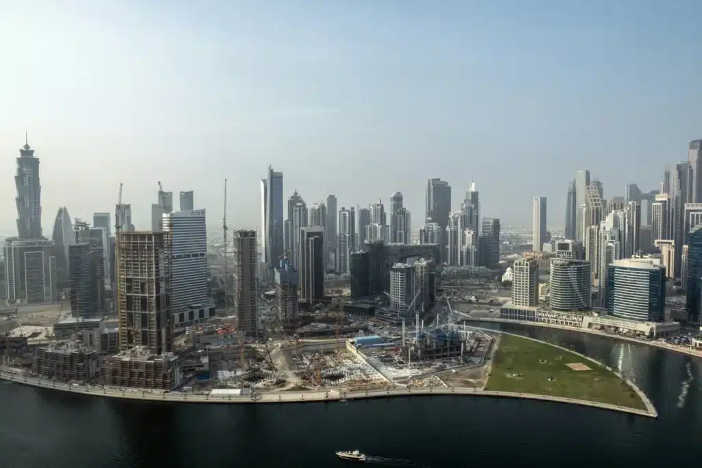 High-angle panoramic view of a dense city skyline featuring numerous skyscrapers, construction sites with cranes, and a large body of water in the foreground under a clear sky.
