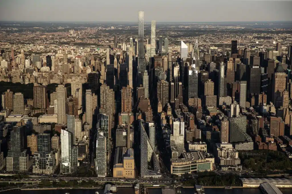 An expansive high-angle aerial photograph of the Midtown Manhattan skyline, featuring modern skyscrapers like Central Park Tower and 432 Park Avenue, with the Hudson River shoreline and a triangular building (VIA 57 West) in the foreground.