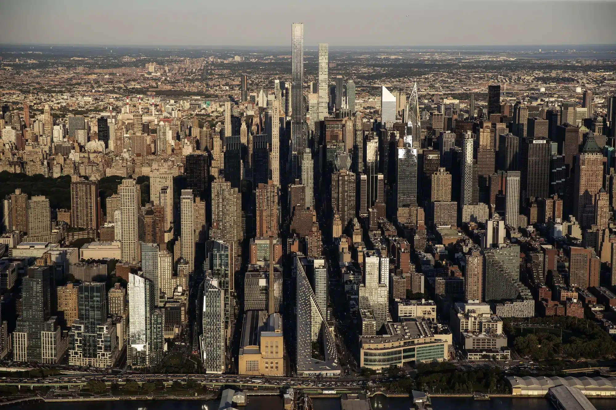 An expansive high-angle aerial photograph of the Midtown Manhattan skyline, featuring modern skyscrapers like Central Park Tower and 432 Park Avenue, with the Hudson River shoreline and a triangular building (VIA 57 West) in the foreground.