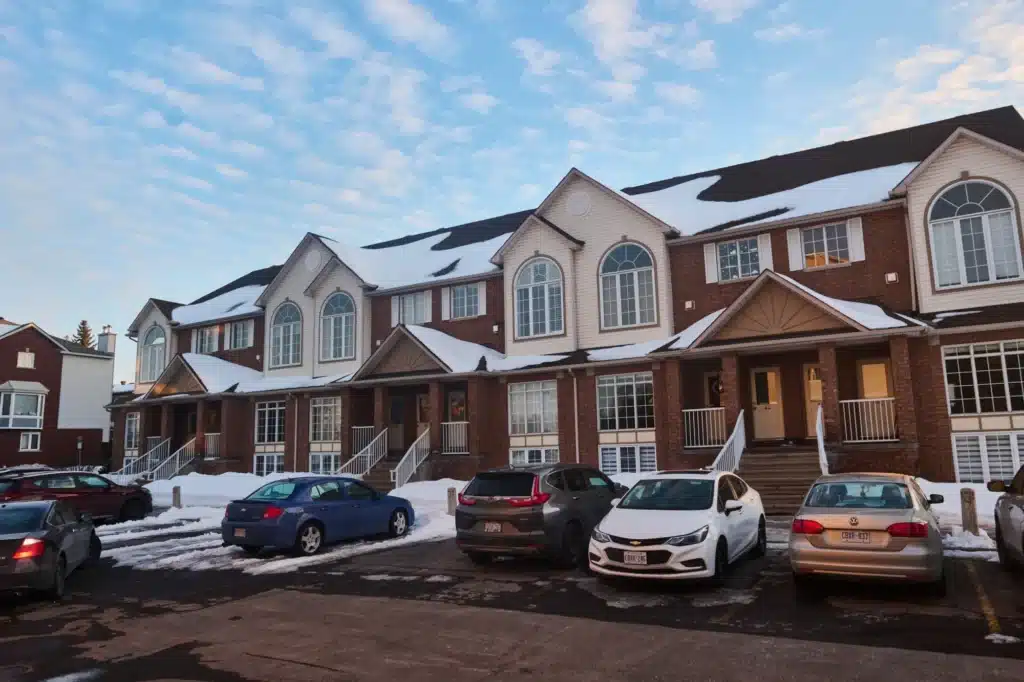 A row of two-story brick townhouses with snow-covered roofs and several cars parked in the front parking lot under a blue sky.