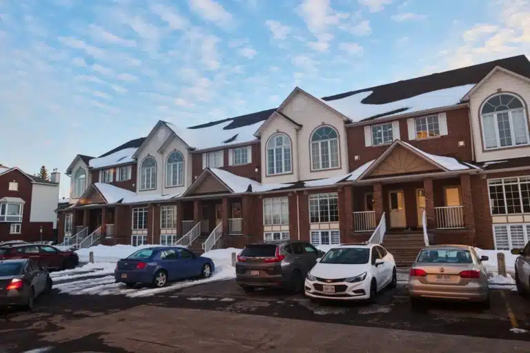 A row of two-story brick townhouses with snow-covered roofs and several cars parked in the front parking lot under a blue sky.