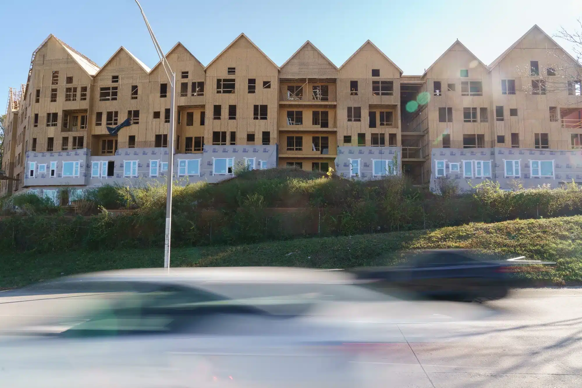 A large multi-story apartment building under construction with exposed wooden framing and several gabled rooflines, situated behind a grassy hill with blurred cars passing in the foreground.