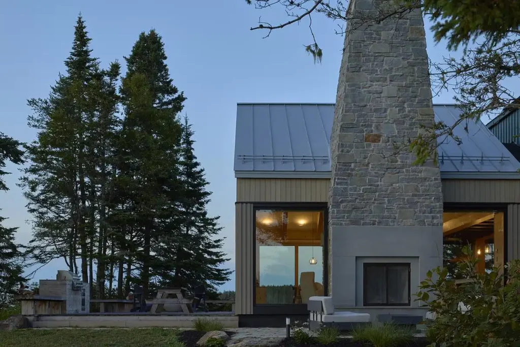 A stone chimney rises beside glowing glass windows against the quiet evening sky.