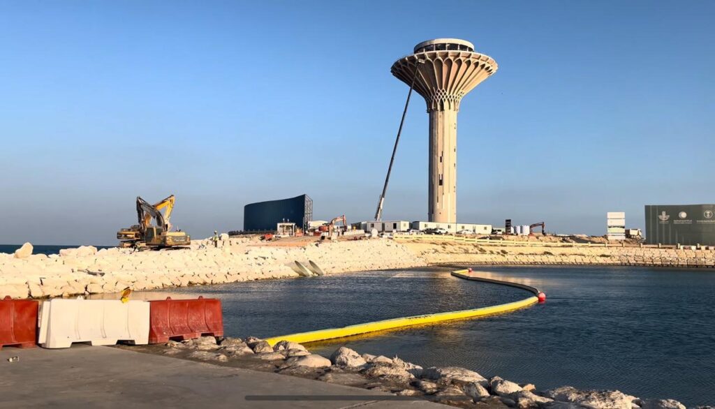 Active construction site on a reclaimed island in Khobar, showing a rising tower core, cranes, and coastal breakwaters.