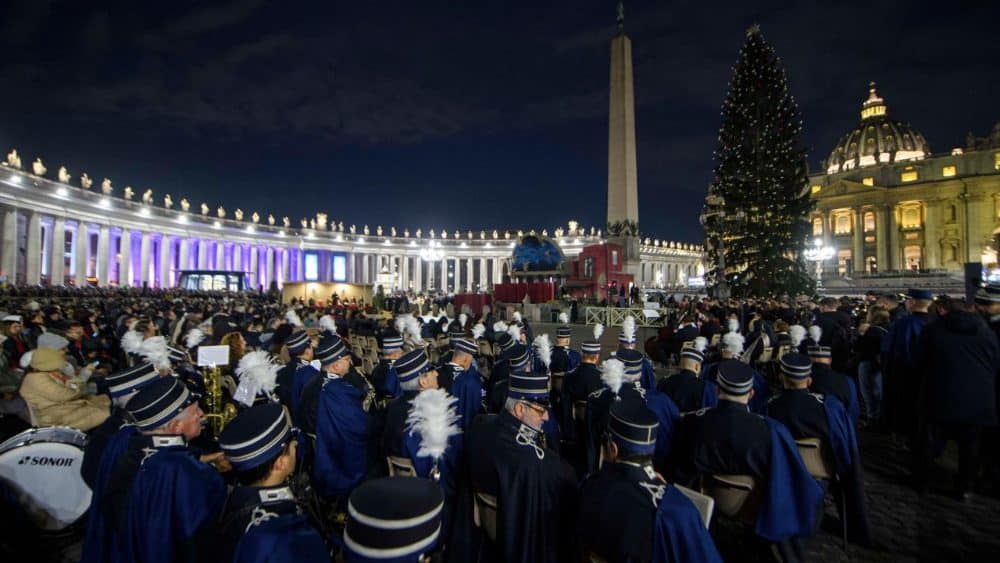 Architecture and temporary installations at St. Peter&rsquo;s Square during Christmas: military band performs beside seasonal tree and Nativity display under Basilica dome.