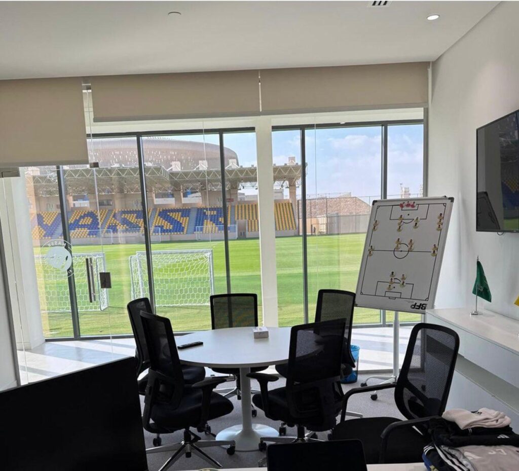 Modern tactical meeting room with round table and strategy board, overlooking Al-Nassr&rsquo;s training pitch and stadium under daylight.
