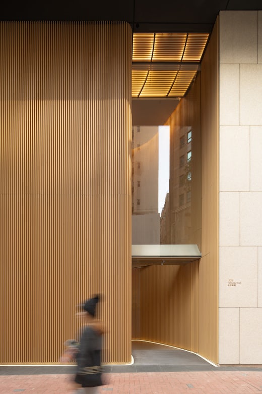 Entrance to a modern office tower in Hong Kong, showcasing vertical wooden fins illuminated from within, glass reflections, and a blurred pedestrian passing by.