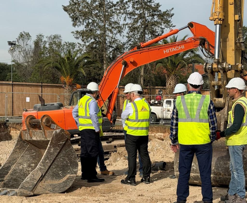 Construction team overseeing site works for the carbon-neutral diplomatic headquarters in Nicosia, standing beside a Hitachi excavator during initial excavation phase.