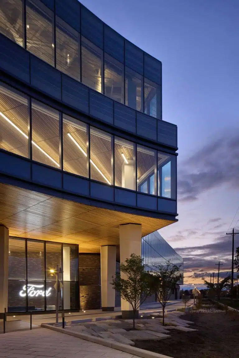 Ford Central Campus Building at dusk, showcasing its modern glass and steel facade with illuminated interior workspaces and a glowing neon "Ford" sign at the entrance, set against a twilight sky in Dearborn, Michigan.