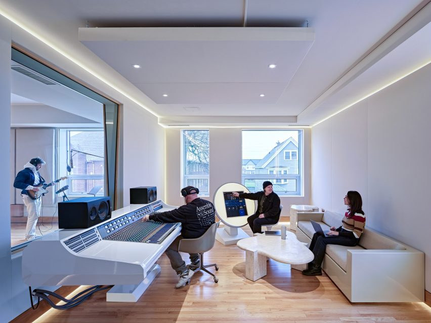 غرفة تحكم حديثة في استوديو تسجيل، تحتوي على مزيج أبيض، يديره شخص بينما يتعاون الآخرون بالقرب منه؛ من خلال الزجاج، يعزف موسيقي على الجيتار في غرفة الأداء، كل ذلك داخل مقر تجميع الصوت في ديترويت ضمن مركز لانترن للفنون.