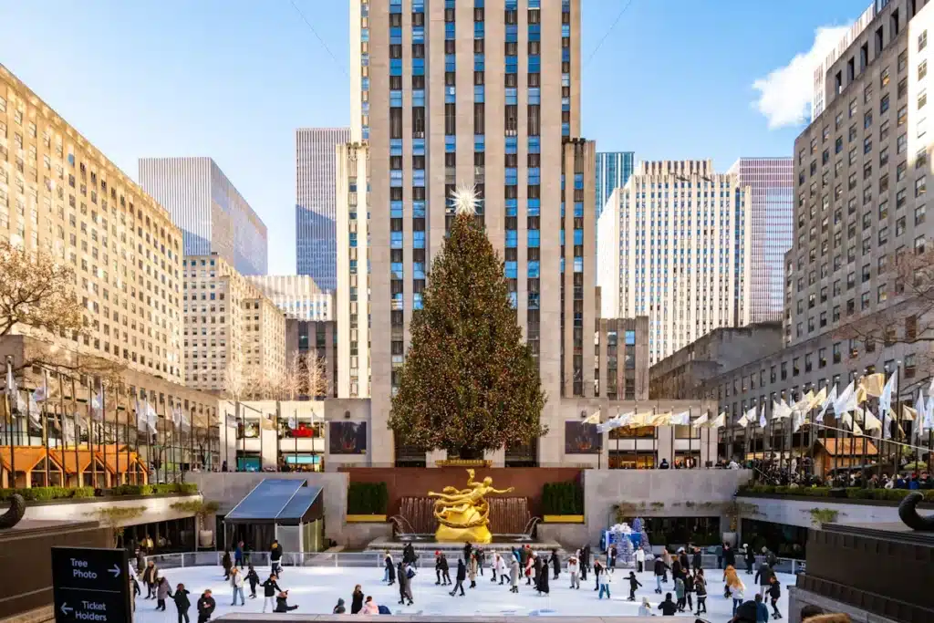 The fully decorated 2025 Rockefeller Center Christmas tree stands tall with a glowing star atop, surrounded by skaters on the ice rink and Art Deco skyscrapers under a clear blue sky.