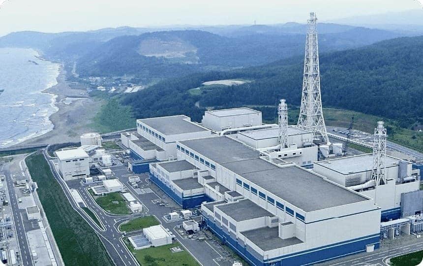 Wide aerial shot of Kashiwazaki-Kariwa Nuclear Power Plant under overcast skies, surrounded by forested hills and Sea of Japan coastline.