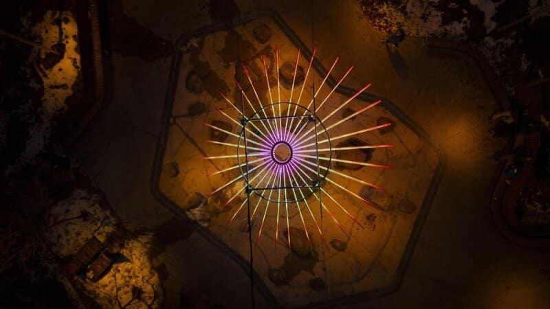 Aerial top-down view of the Un Brin d&rsquo;Soleil light installation at night, showing its radial LED structure glowing in purple and orange hues above a snow-dusted urban plaza.