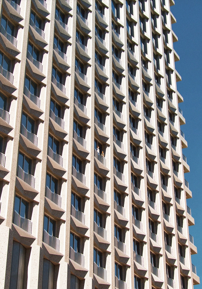 A close-up view of a high-rise building’s facade with repeating angular balconies under a clear blue sky, highlighting its rhythmic architectural pattern and material texture.