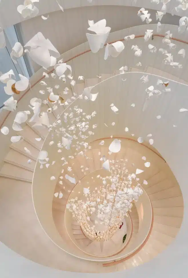 Interior view of the Dior Beijing flagship store, showing a white spiral staircase with suspended petal-like sculptural elements against a light wood ceiling.