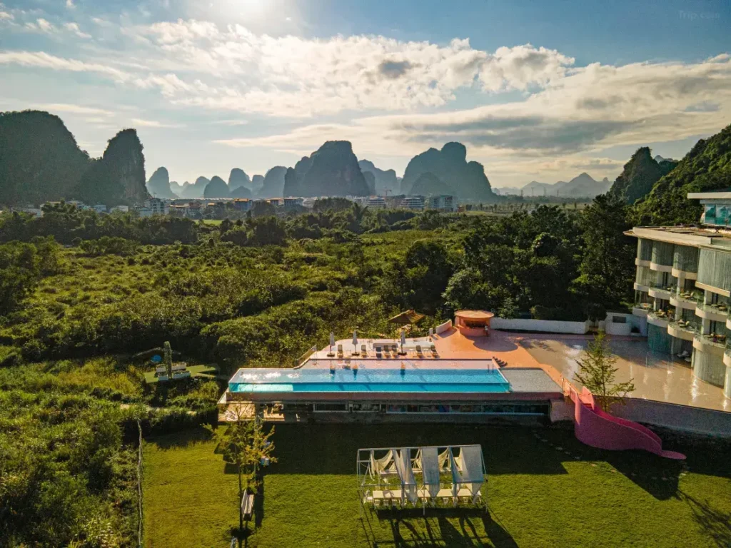 Aerial view of LS Hotel in Guilin, showcasing its rooftop pool and pink slide against a backdrop of lush greenery and iconic karst mountains under a partly cloudy sky.